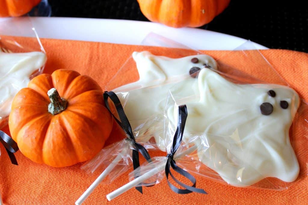 Two white chocolate ghost pops on an orange napkin with a mini pumpkin and black ribbon.