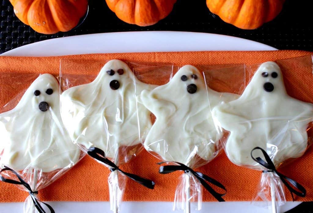 Overhead photo of 4 White Chocolate Ghost Pops on a white plate with an orange napkin and 3 small pumpkins.