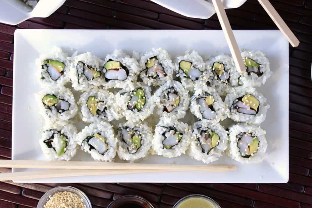 Overhead picture Sushi Rolls with Avocado and Shrimp on a long white plate with sesame seeds and chopsticks.