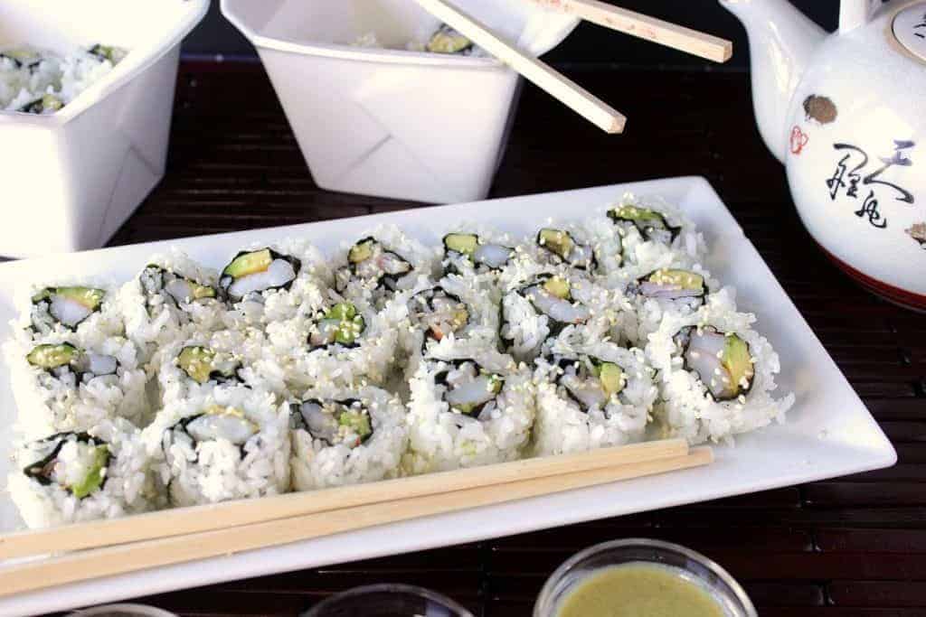 Homemade avocado shrimp sushi rolls on a white plate with chopsticks and sauces.