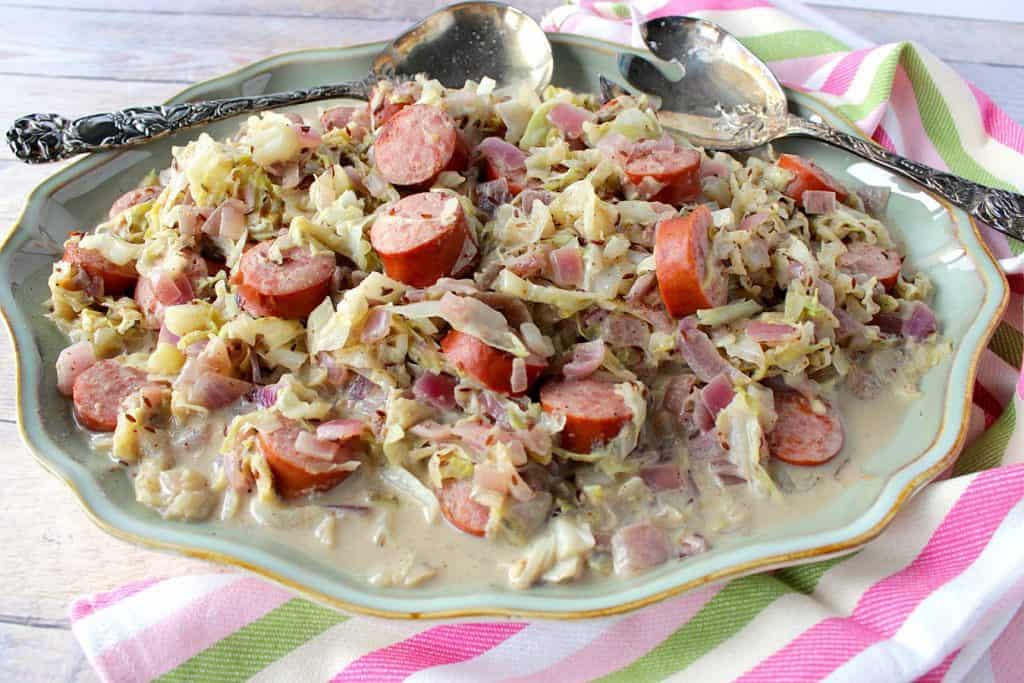 Horizontal photo of a platter filled with creamed cabbage with caraway and sausage with a pink, green, and white striped napkin and serving spoons.