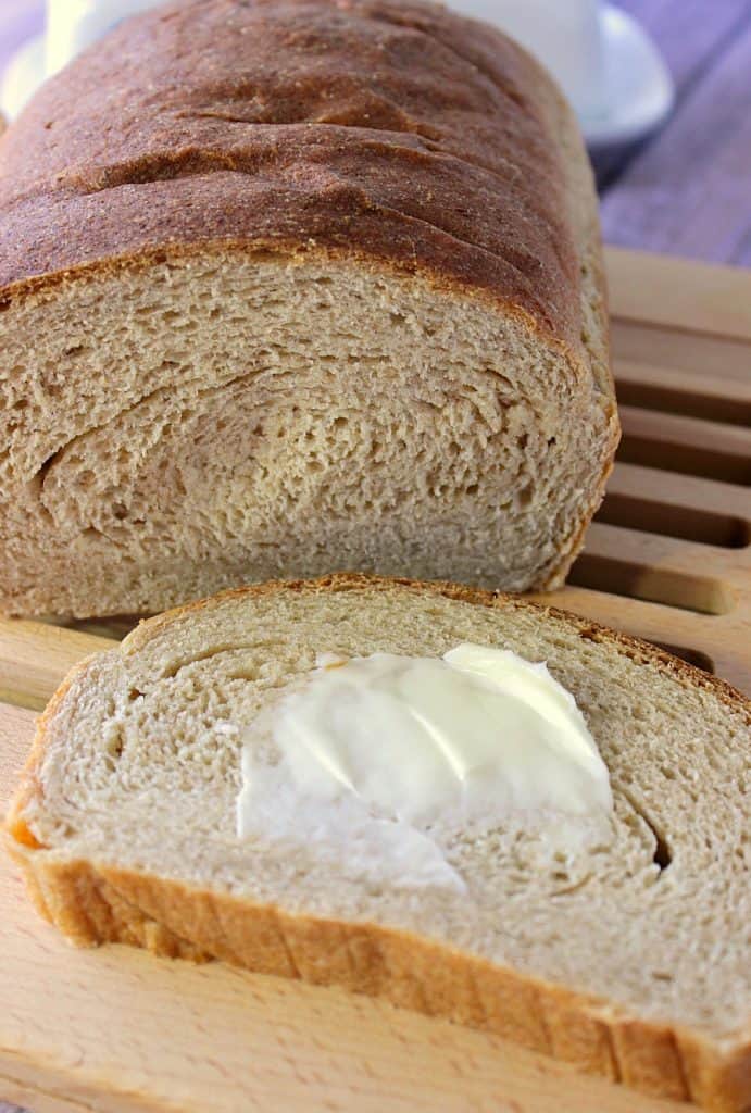 A vertical close up photo of a loaf of Whole Wheat Honey Ricotta Bread with a slice taken out and smeared with butter.
