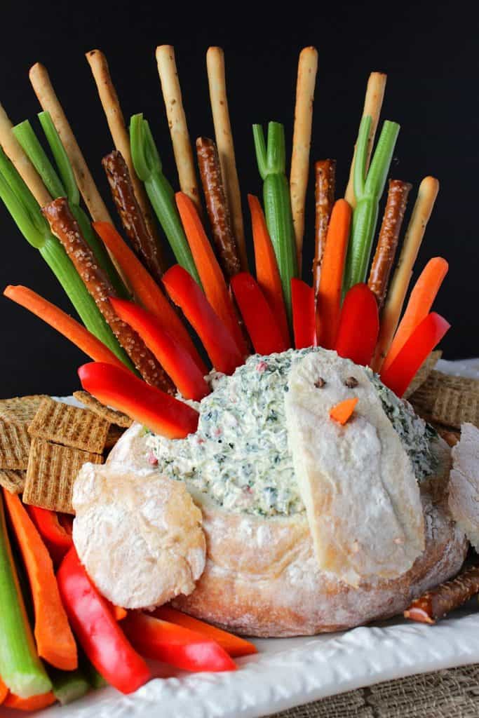 A vertical photo of a colorful turkey bread bowl filled with dip and with veggie tail feathers.