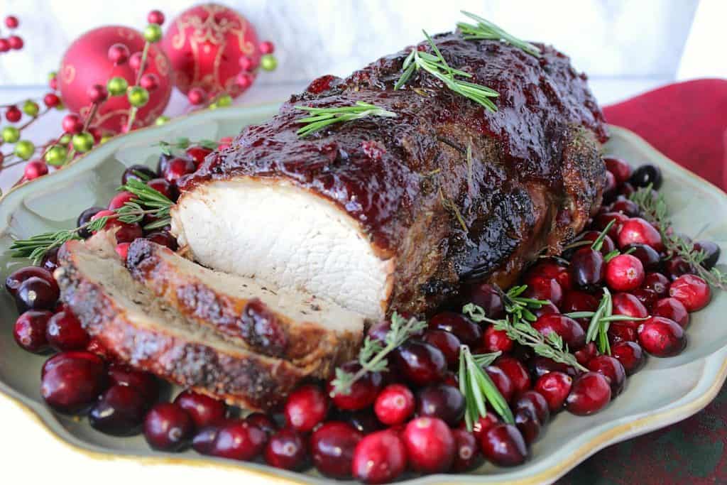 A horizontal photo of a holiday sliced pork roast on a platter with cranberries and rosemary.