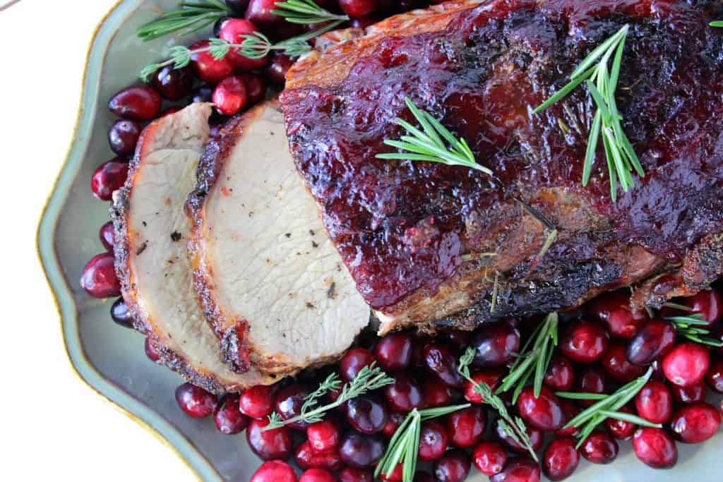  An overhead photo of a sliced Roast Pork with Cranberry Relish on a platter with fresh cranberries and fresh herbs.
