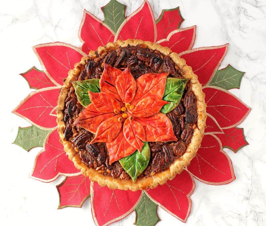 An overhead horizontal photo of a pecan pie with a painted poinsettia pie crust on a red and green poinsettia place mat.