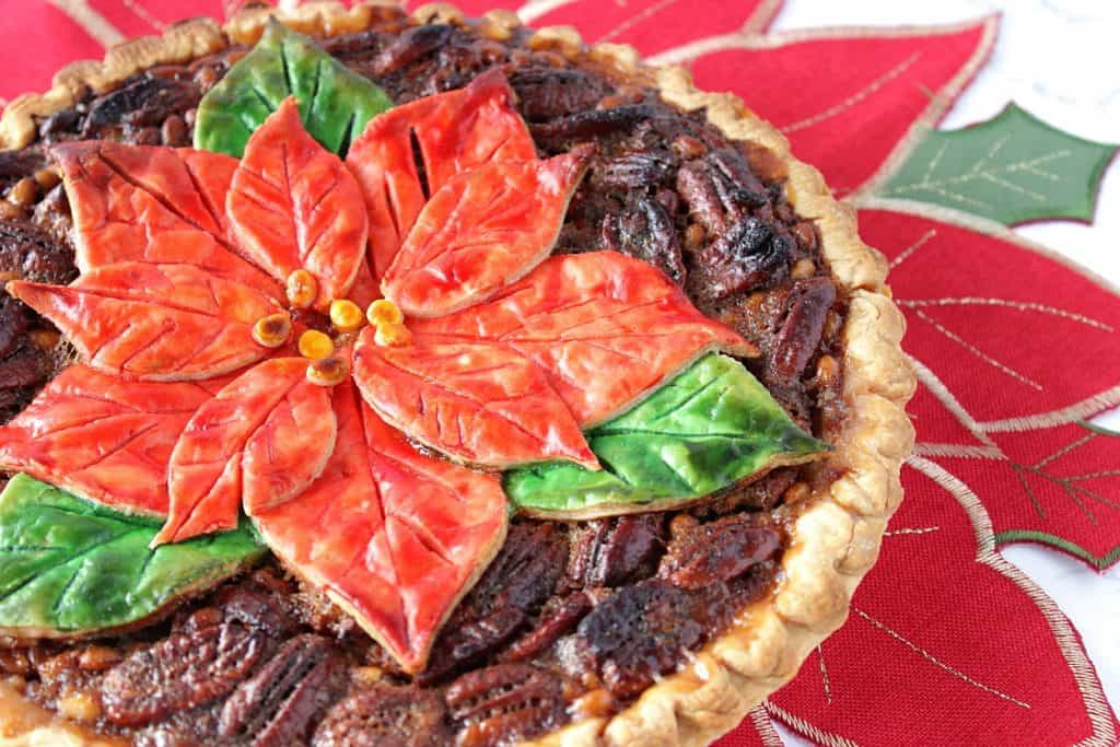 An offset horizontal closeup photo of a pretty poinsettia painted pecan pie on a poinsettia place mat.