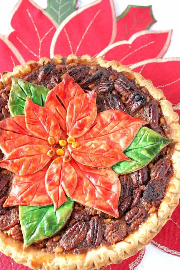 A vertical offside overhead and closeup photo of a pecan pie with a painted poinsettia crust on top.