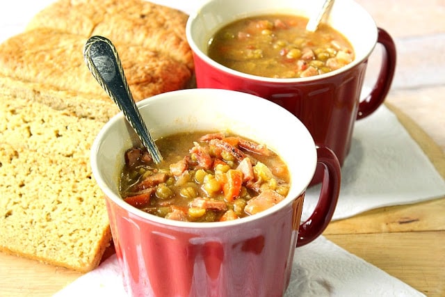 Two mugs of Split Pea Soup with a loaf of bread and two spoons.