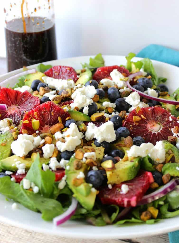 A vertical image of a white salad bowl filled with Blood Orange Salad with Blueberries and Feta cheese along with a bottle of dressing in the background.