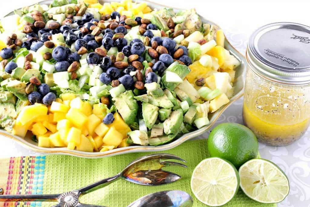 A horizontal photo of an avocado and mango salad on a platter with limes, tongs, and a jar of homemade vinaigrette on the side.