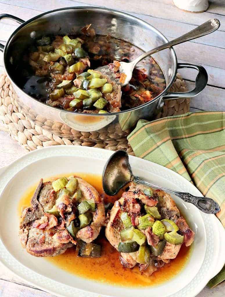 Two pork chops on a platter with dill pickles and a pan in the background