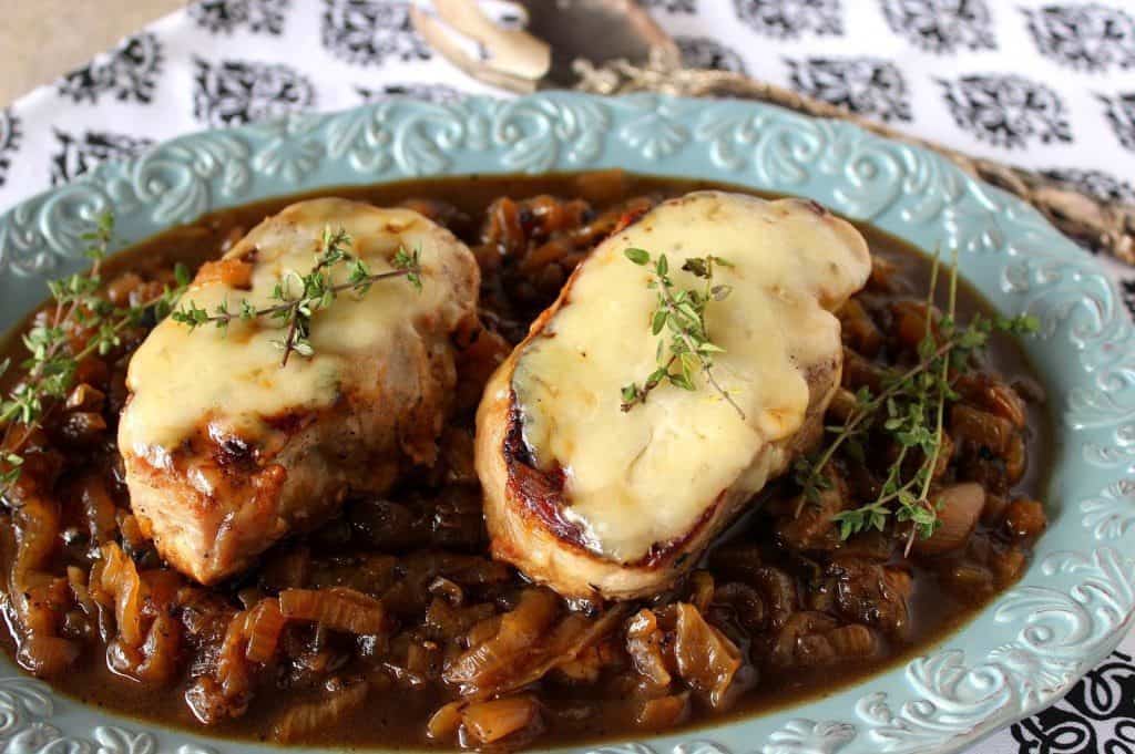 Two juicy French onion pork chops on a pretty blue plate with cheese, caramelized onions and fresh thyme sprigs.