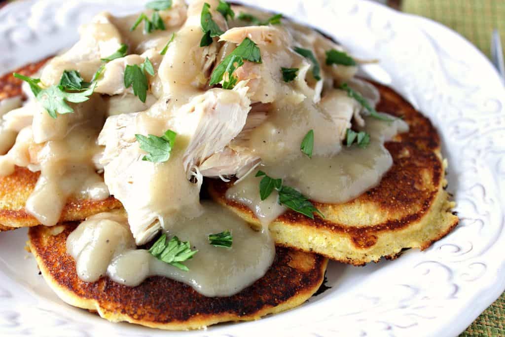 A closeup horizontal photo of a plate filled with corn cakes topped with shredded chicken and a buttery maple gravy.