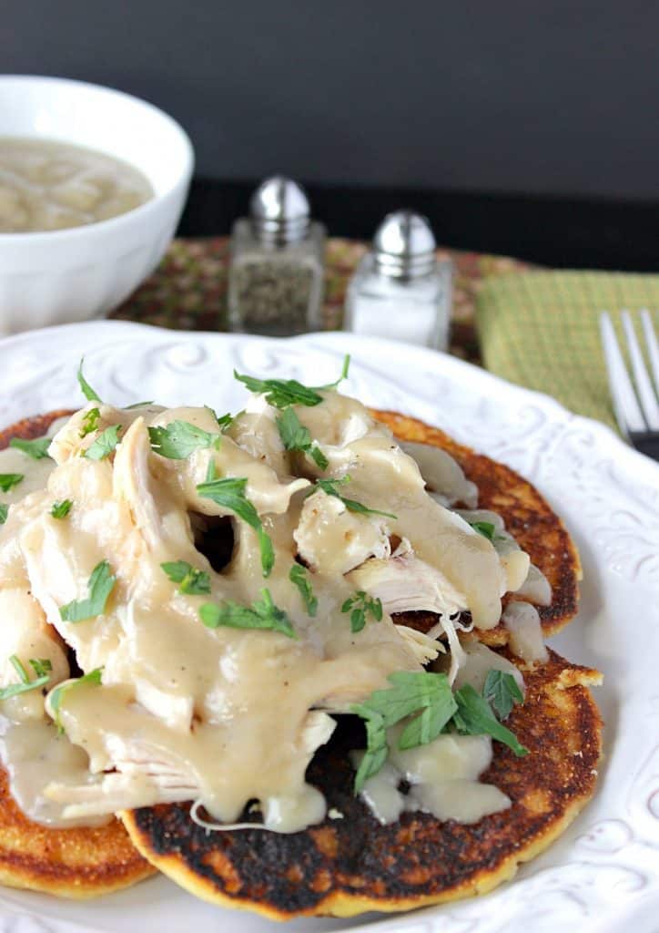 A vertical closeup photo of a bunch of corn cakes on a plate topped with chicken and buttery maple gravy