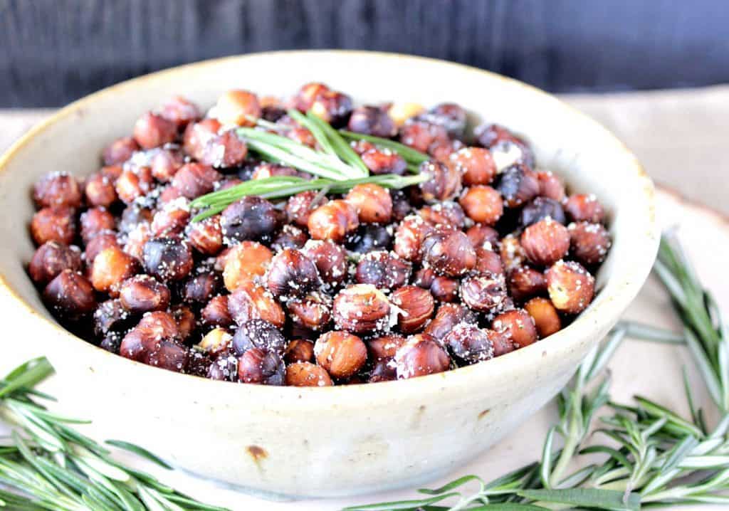 A white bowl filled with Roasted Rosemary Hazelnuts with fresh rosemary for garnish