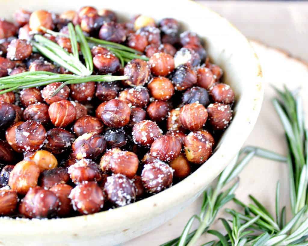 A closeup photo of a bowl filled to the rim with Skillet Roasted Rosemary Hazelnuts along with fresh sprigs of rosemary as garnish