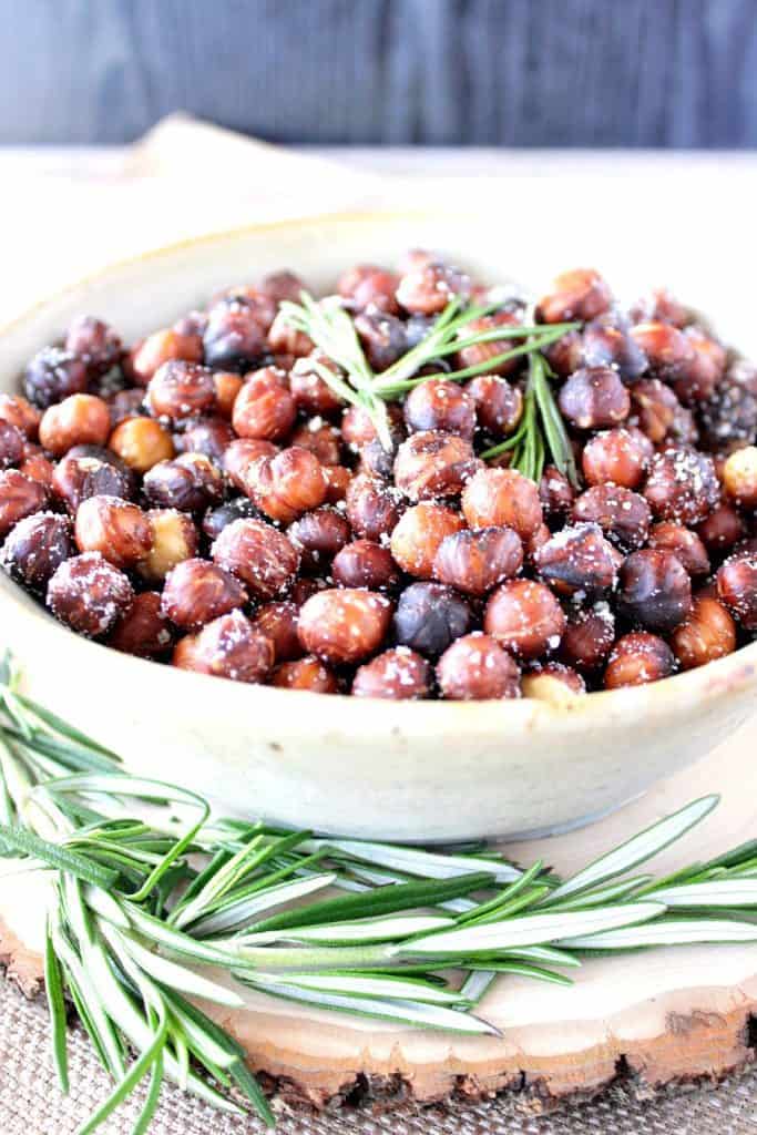 A closeup photo of a bowl filled with Skillet Roasted Rosemary Hazelnuts with fresh rosemary as garnish 