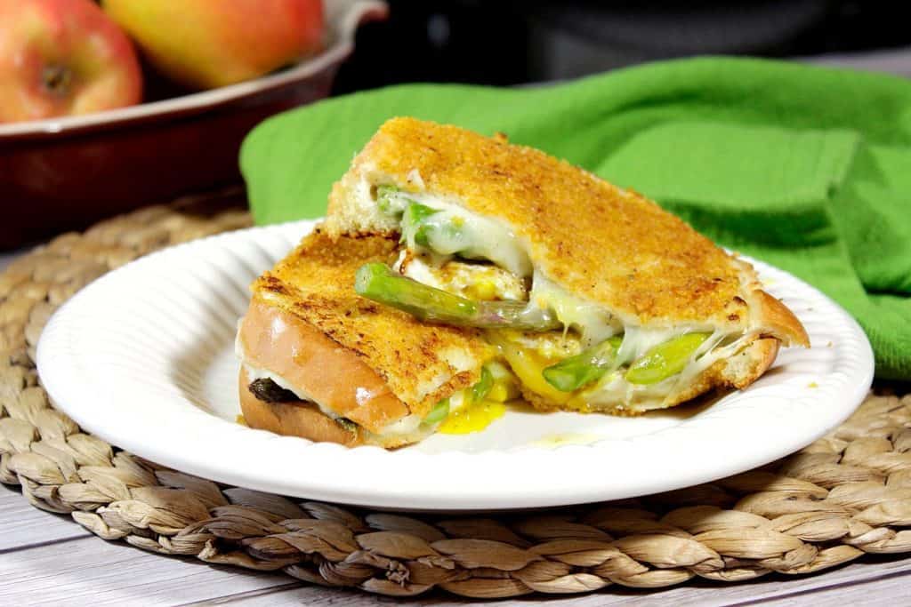 Grilled Cheese Breakfast Sandwich with Egg, Asparagus, and Provolone Cheese. Grilled cheese breakfast sandwich on a white plate with a green napkin and apples in the background.