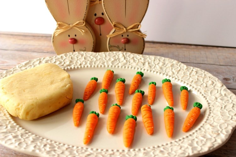 Homemade marzipan candy carrots on a white plate with a cute wooden bunny face plaque in the background.