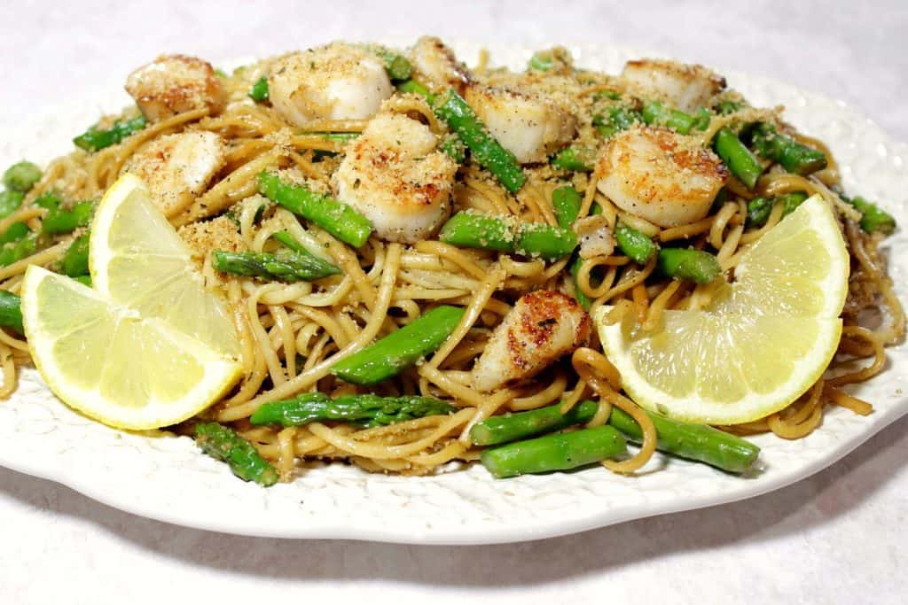 A platter filled with seared scallops, linguine, asparagus, and garnished with lemon slices and breadcrumbs.