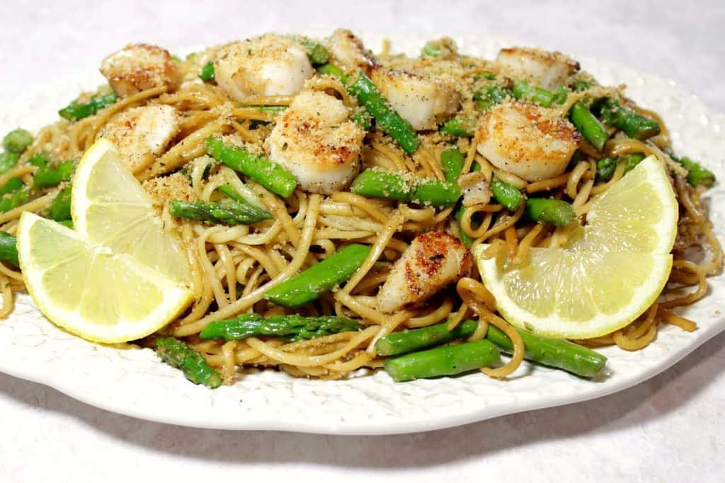 Sweet Caramelized Seared Scallops are what dreams are made of. Especially when served with linguine, asparagus and tossed with buttered breadcrumbs! | Kudos Kitchen by Renee A platter filled with seared scallops, linguine, asparagus, and garnished with lemon slices and breadcrumbs.