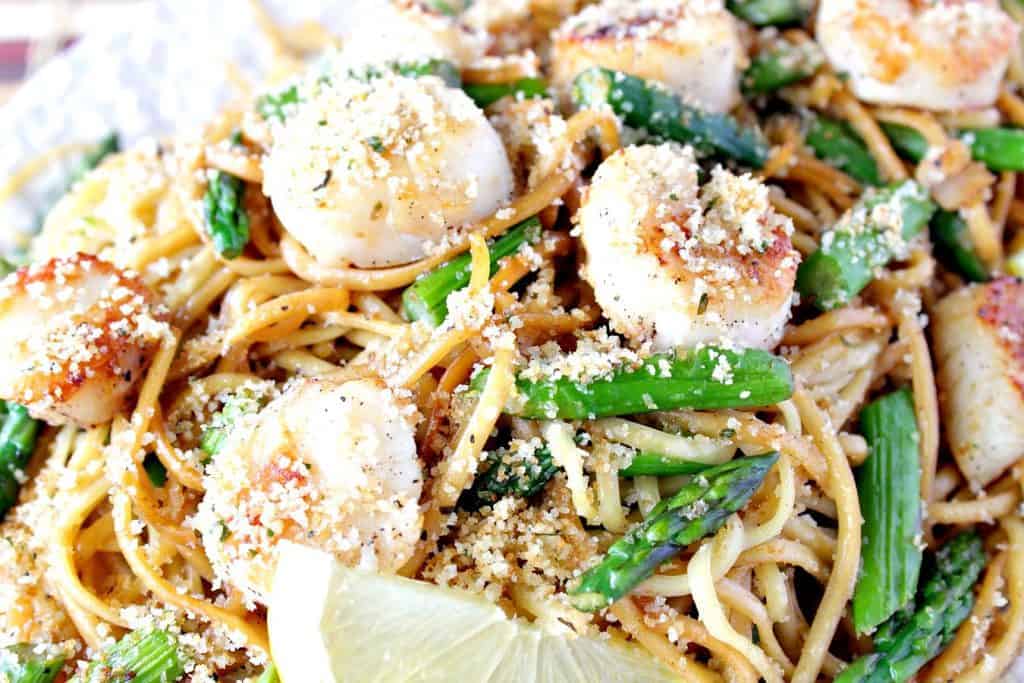 A closeup overhead photo of scallops with linguine and asparagus, breadcrumbs and lemon slices.