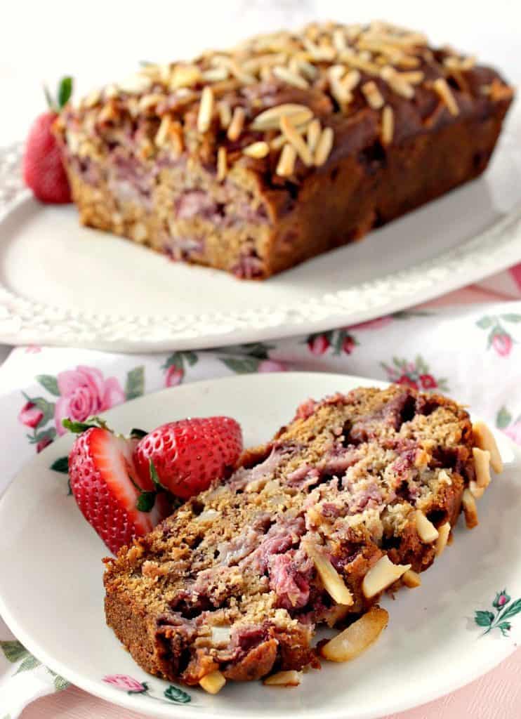A vertical closeup photo of a slice of strawberry banana bread on a plate and the entire loaf in the background.