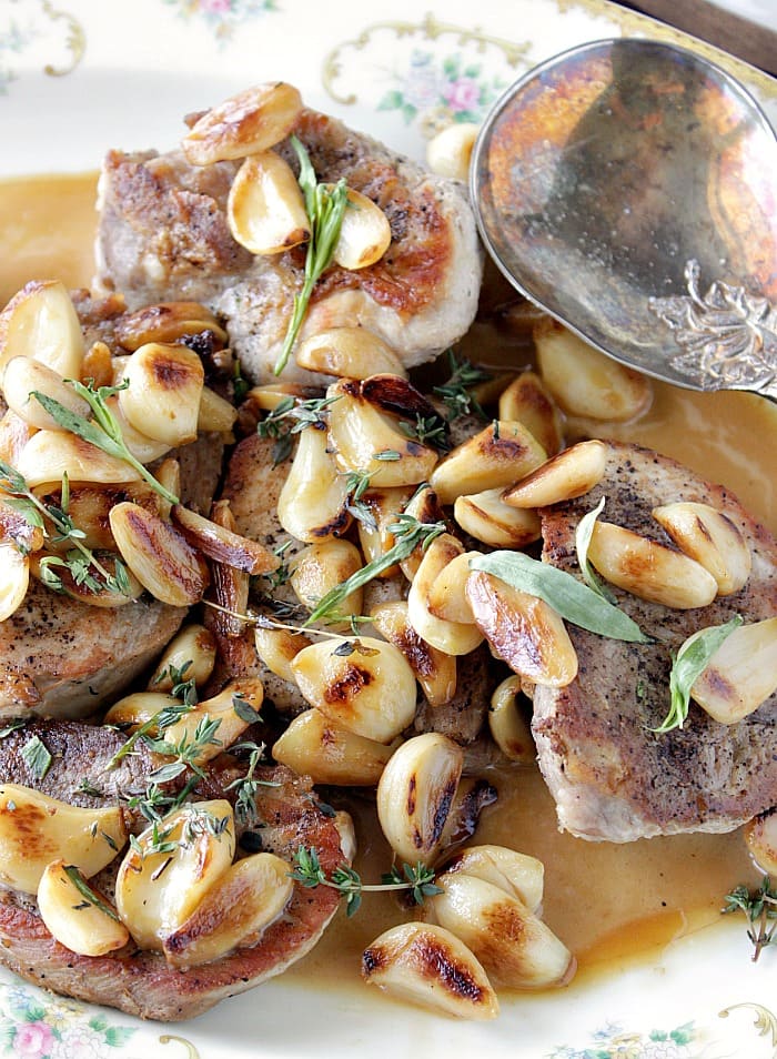 Vertical closeup photo of lots of browned garlic cloves over seasoned pork chops with fresh herbs.
