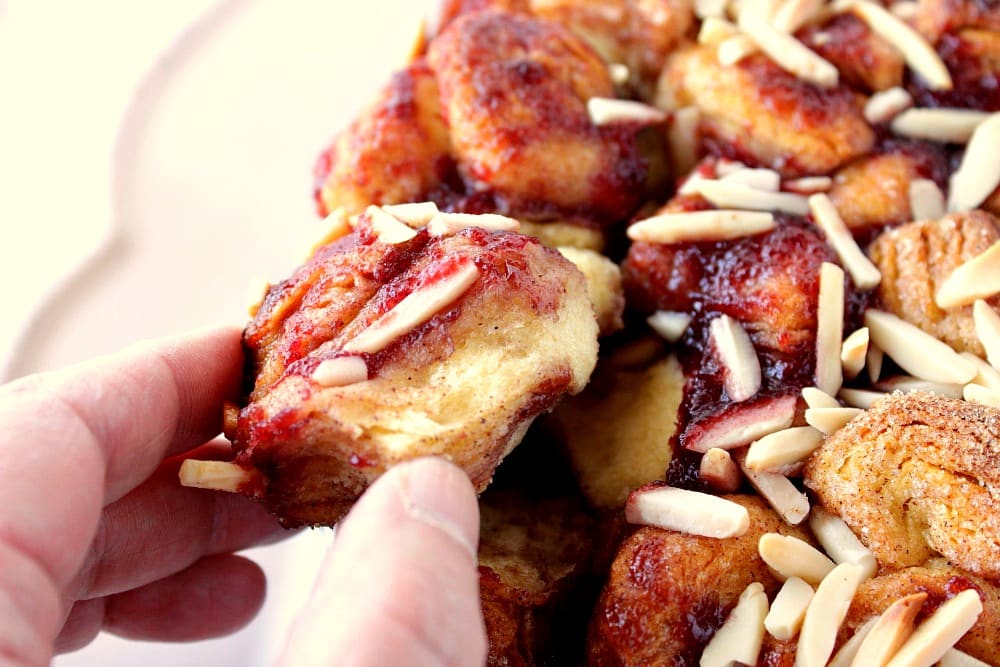 A closeup photo of a hand pulling off a piece of raspberry spice monkey bread with almonds.