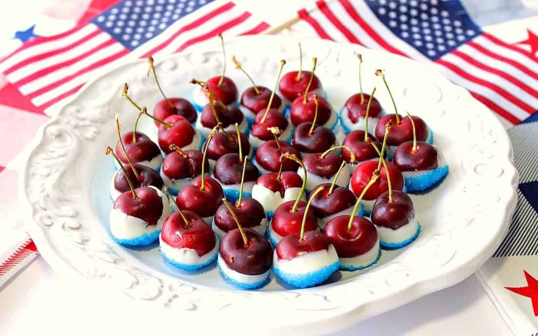 A white oval plate filled with red white and blue sugared cherries with American flags in the background.
