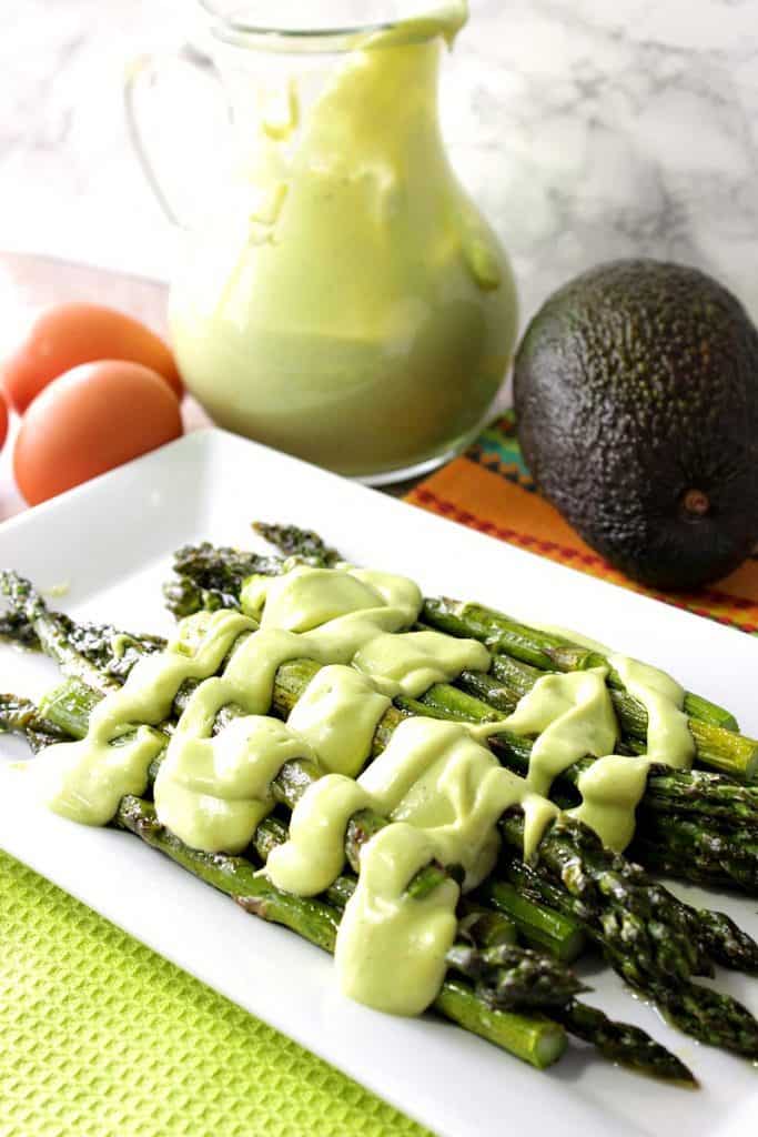 Avocado hollandaise sauce poured over a plate of asparagus. Eggs and an avocado are in the background with additional sauce.