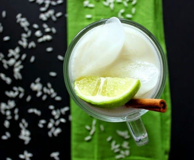 An overhead closeup image of a mug of homemade horchata with ice, a cinnamon stick, wedge of lime, and grains of rice on a black and bright green background.