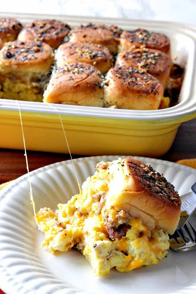 Vertical photo of a plate with a scrambled egg breakfast slider with cheese, and a baking dish filled with the sliders in the background.