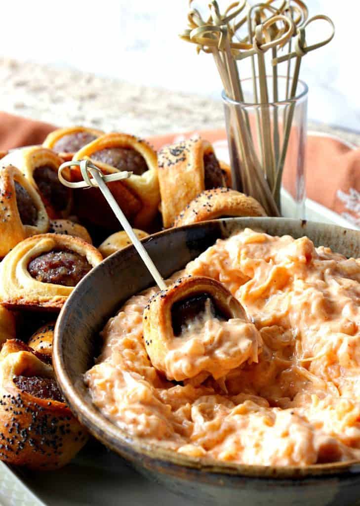 A closeup vertical photo of a bratwurst appetizer bite dipped in a bowl of sauerkraut dipping sauce with a cocktail pick.