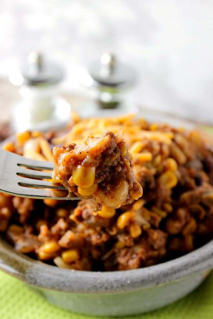 Vertical closeup image of a bowl of chili mac on a fork with macaroni, corn, ground beef and salt and pepper shakers in the background.