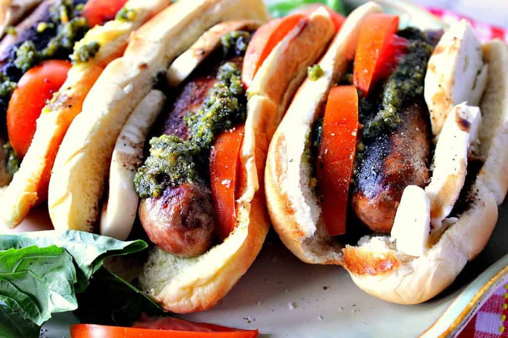 Three caprese sausage sandwiches on a platter with tomatoes, mozzarella cheese, and basil pesto.