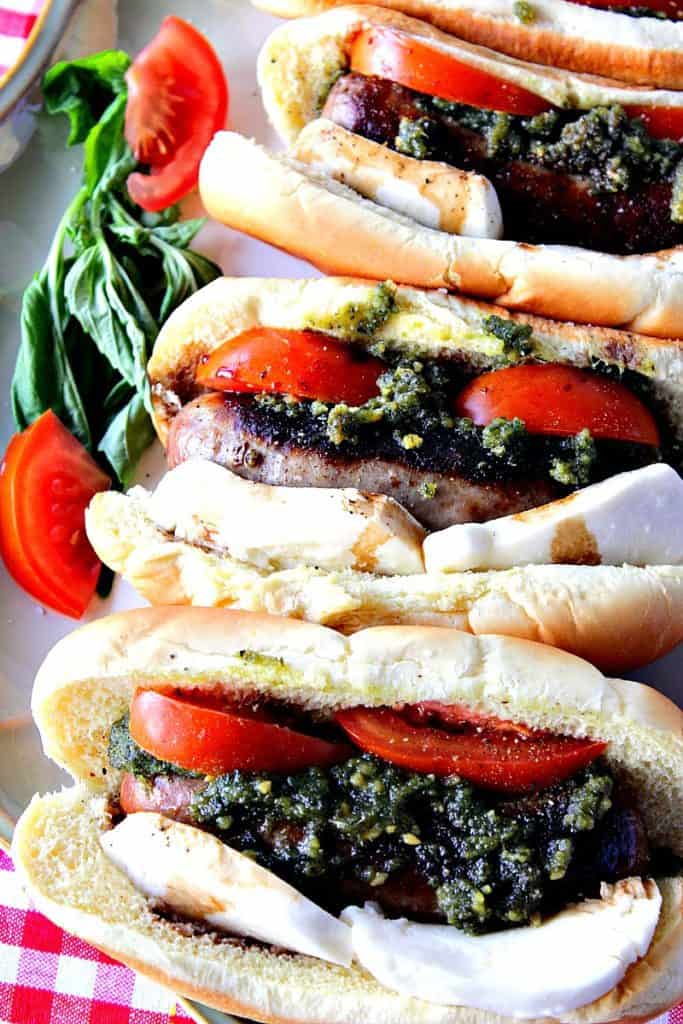 Vertical image of three Caprese Sausage sandwiches with basil pesto, tomato slices, fresh mozzarella cheese, on sausage buns.