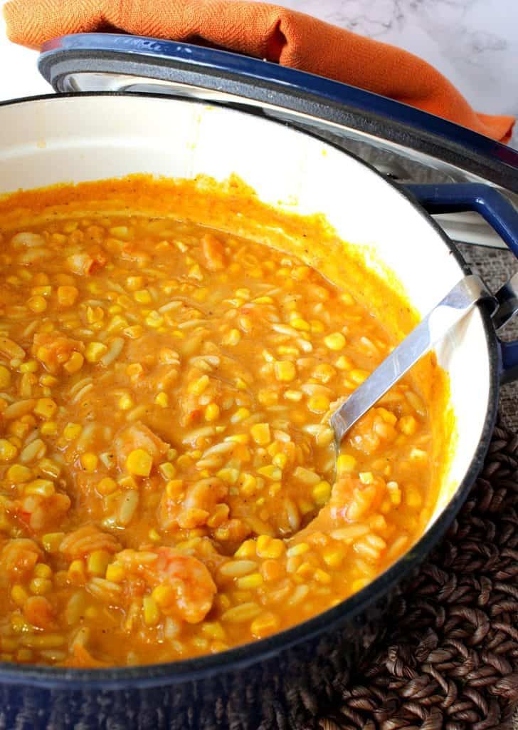 A vertical closeup photo of a pot of pumpkin corn chowder with shrimp with a ladle and an orange napkin.