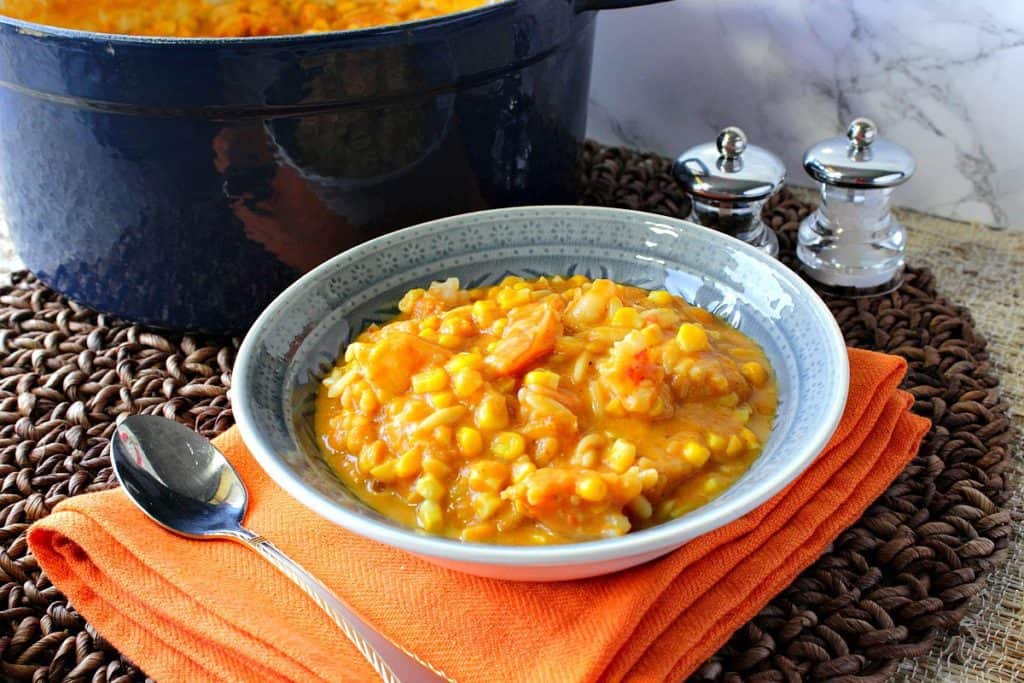 A blue bowl filled with corn chowder with pumpkin and shrimp with a spoon and an orange napkin.