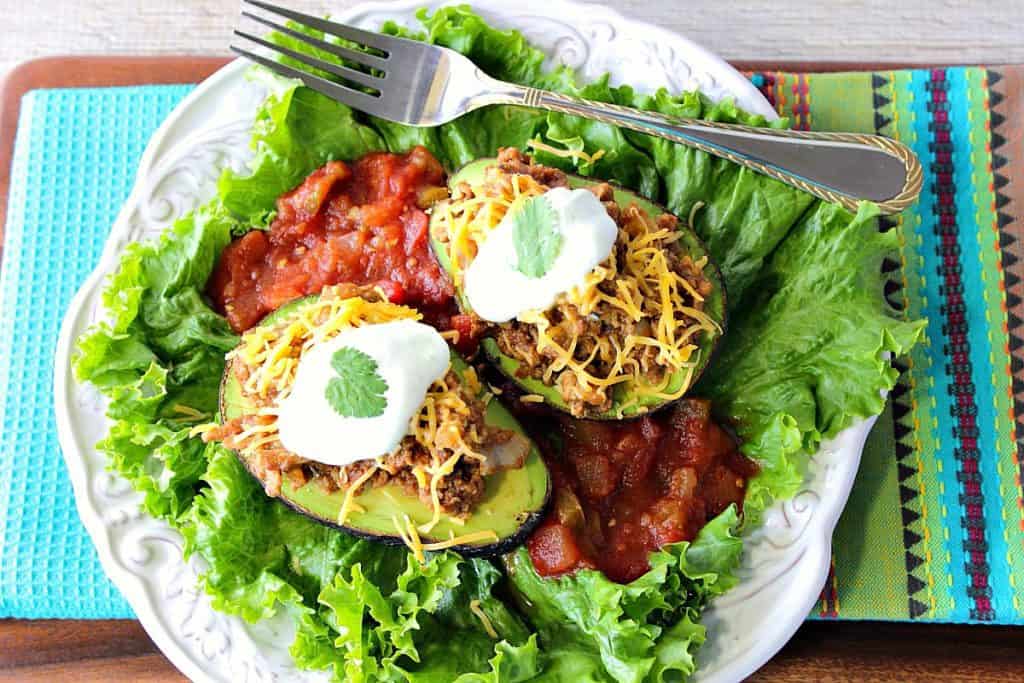 Two stuffed avocados on a plate with lettuce, salsa, shredded cheese, sour cream, a fork, and a bright blue napkin.