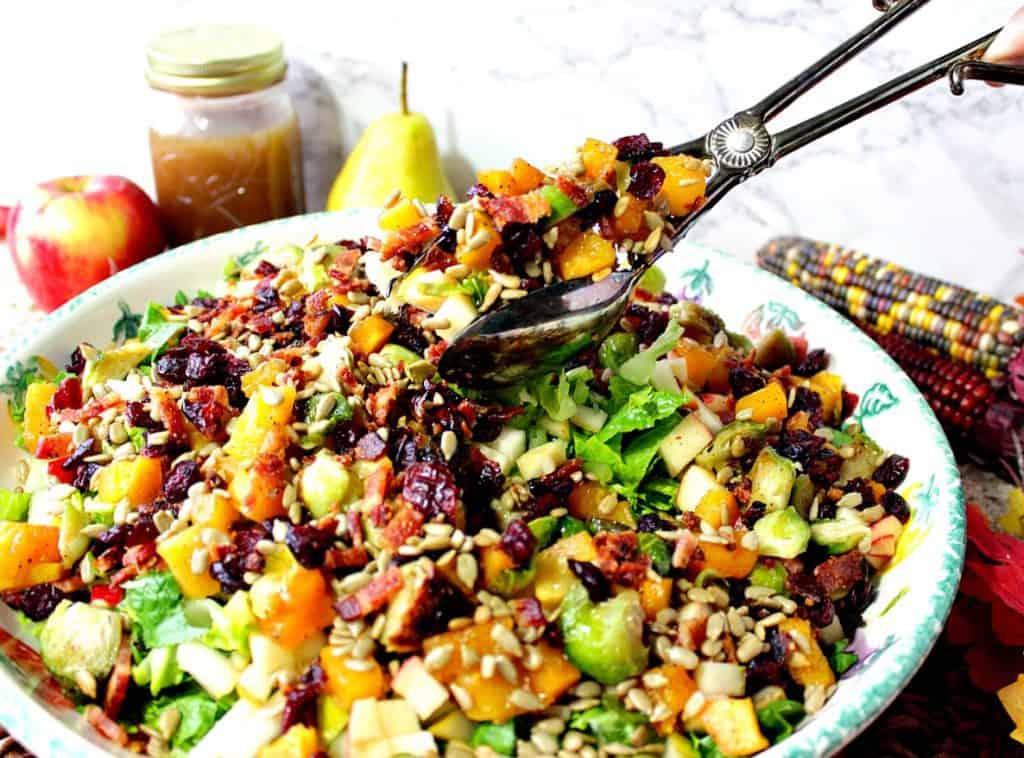 Salad tongs holding a large portion of fall chopped salad with Brussels sprouts, dried cranberries, and bacon.