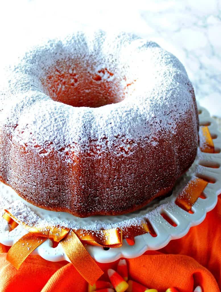 A citrus bundt cake with confectioners sugar dusting on a cake stand with an orange ribbon.