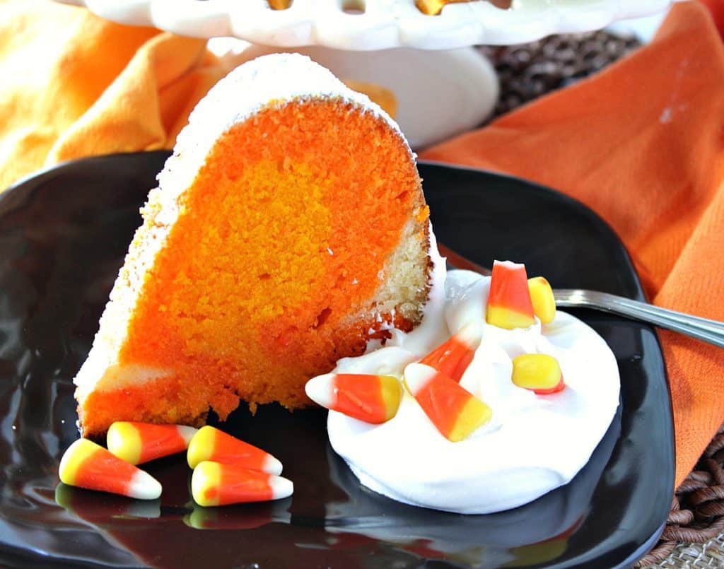 A horizontal photo of a slice of candy corn colored pound cake on a black plate with whipped cream and candy corn.