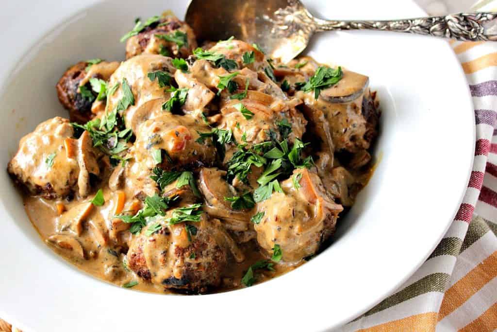 A large white bowl filled with German meatballs with caraway mushroom cream sauce, parsley, and a large serving spoon.