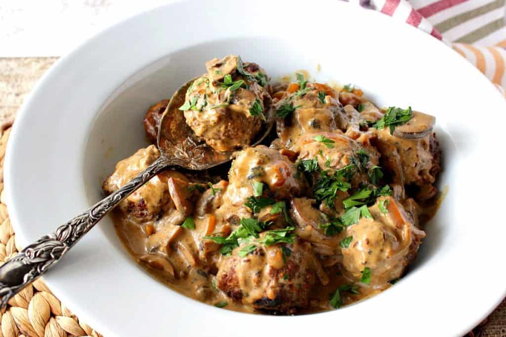 A white bowl filled with German meatballs, cream sauce, parsley, and a spoon.