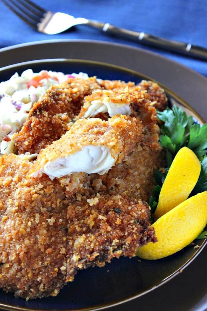 A vertical closeup image of breaded fried tilapia on a plate with lemon wedges, parsley, and coleslaw.