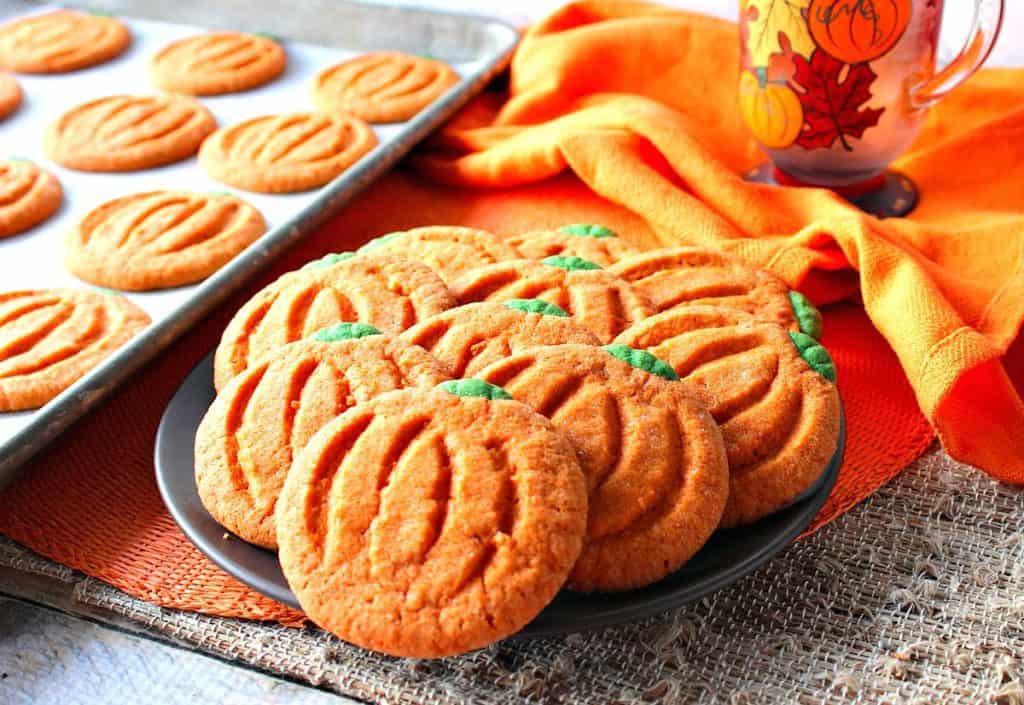 A horizontal image of a plate filled with orange and green pumpkin shaped sugar cookies with an orange napkin and a textured place mat.
