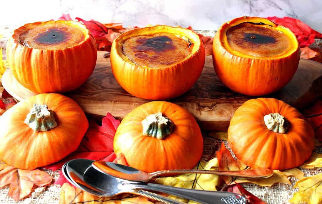 Three pumpkin creme brulee baked in real pumpkins lined up on a board with autumn leaves and pumpkin tops.