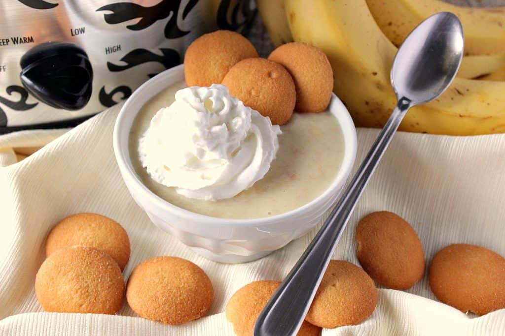 Slow Cooker Banana Coconut Pudding A bowl of banana coconut pudding with vanilla wafer cookies, a spoon, and a dollop of whipped cream.